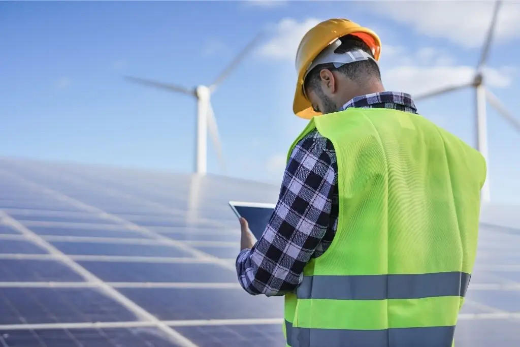 Ein Techniker in Sicherheitsweste und Helm inspiziert Solarmodule mit Windrädern im Hintergrund und hält dabei ein Tablet.