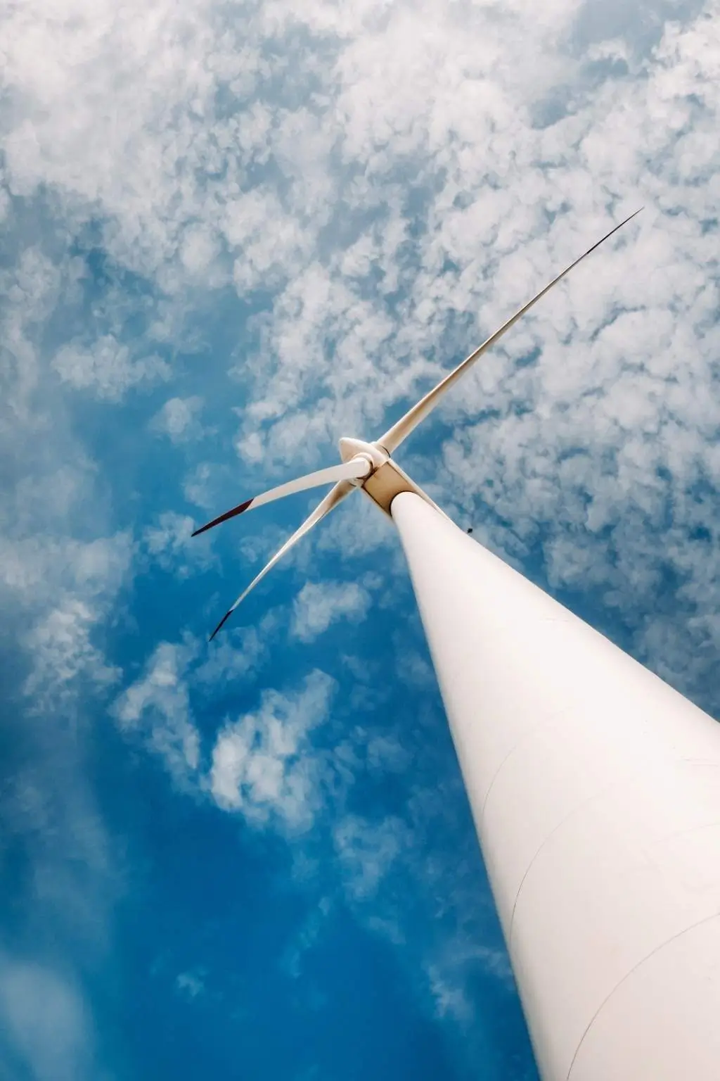 Nahaufnahme einer Windturbine aus der Froschperspektive vor einem wolkigen blauen Himmel.
